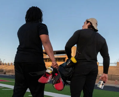 Two people on a football field carrying athletic equipment.